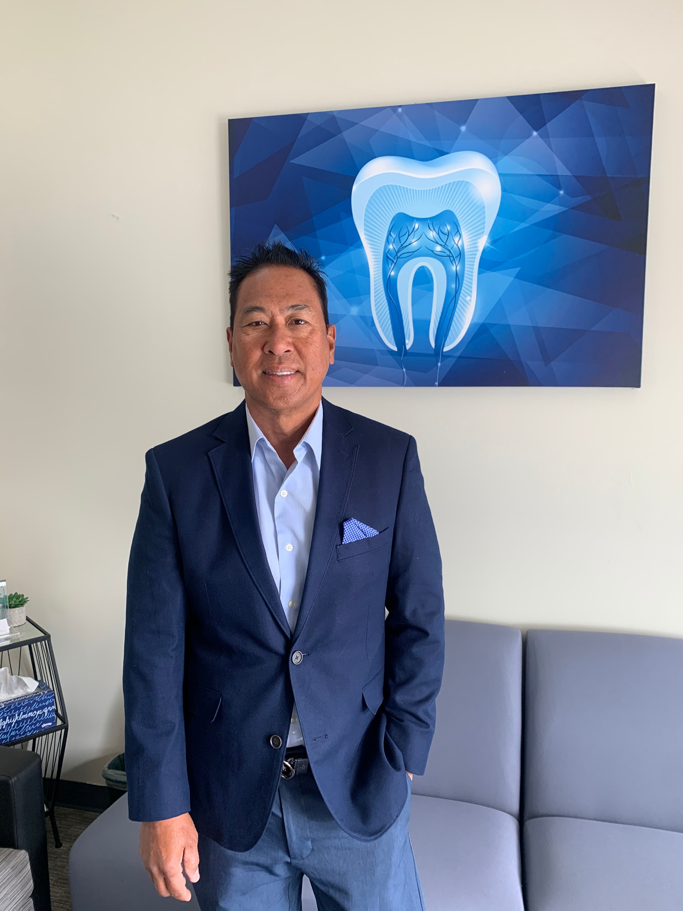 Dr. Ron E. Quibilan standing in the Sunnyvale office waiting area, wearing a navy blazer in front of the blue tooth artwork.