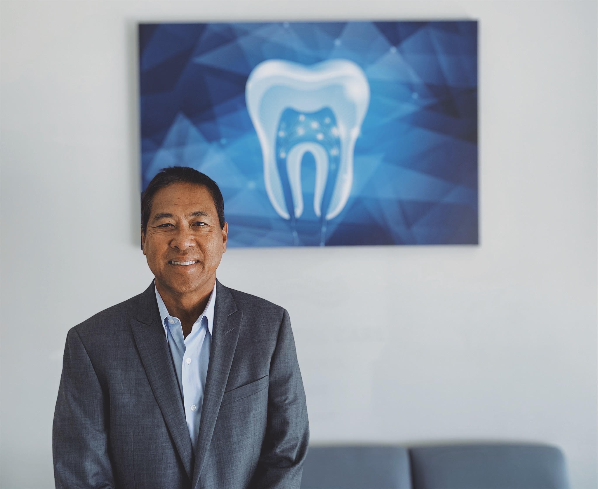 Dr. Ron E. Quibilan, DDS, smiling in a gray suit in front of dental artwork in the waiting area.